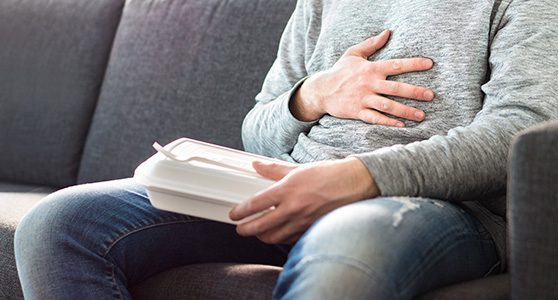 Uma fotografia de um homem sentado no sofá com uma marmitex em uma mão e a outra na barriga com intoxicação alimentar representando a necessidade do seguro responsabilidade civil produtos.