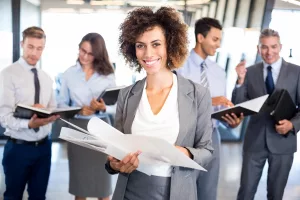 Mulher de roupa social sorrindo a frente de restante de equipe corporativa